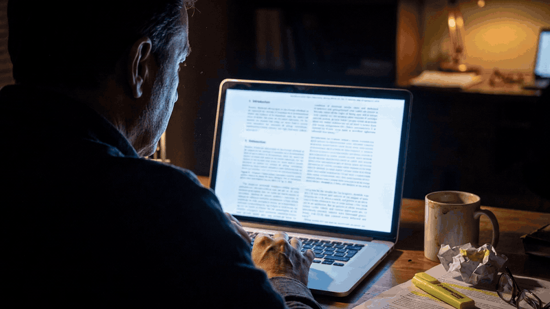 A man reading research documents on a laptop at a desk in the evening